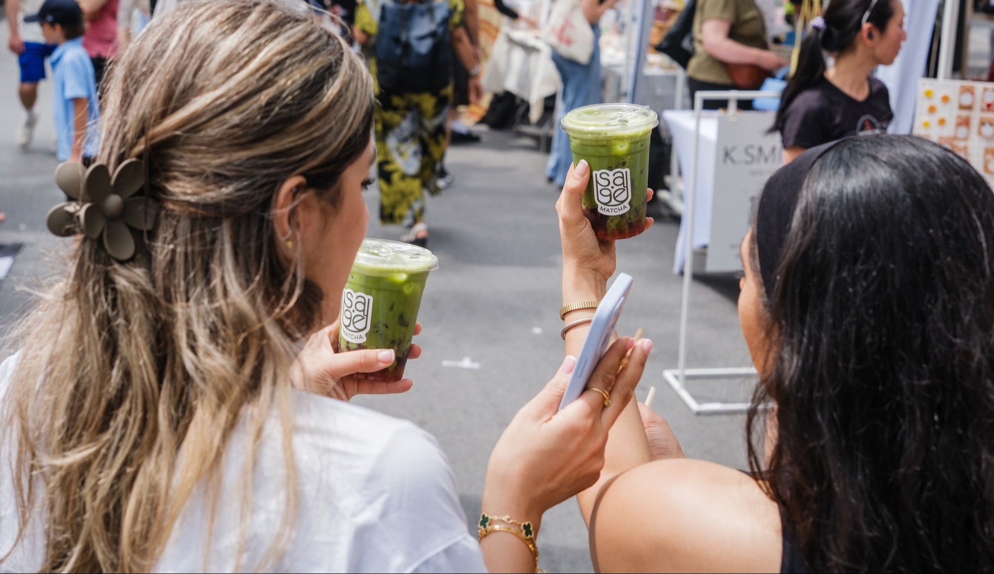 Two women holding green drinks and a phone in an outdoor setting with people and tents in the background.
